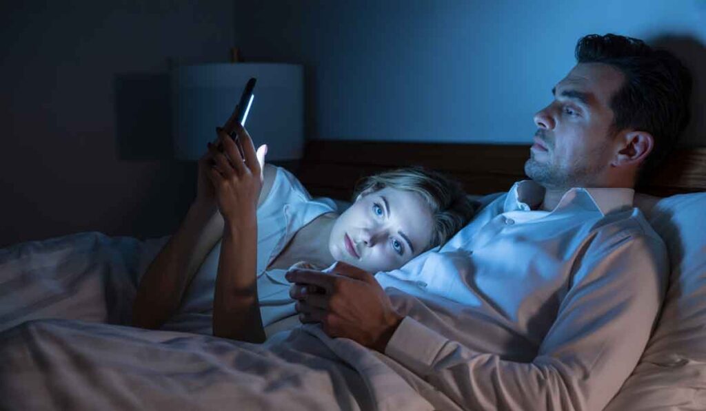 A man and woman lie in bed at night, both looking at their smartphones. The woman rests her head on the man's shoulder, illuminated by the light from their screens.